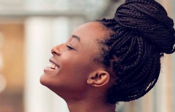 mulher negra de lado sorrindo e levando a cabeça com penteado de tranças em formato de coque
