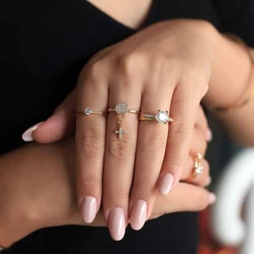 Mão feminina com unhas feitas, anel de cruz no dedo do meio e anéis solitários nos dois dedos ao lado