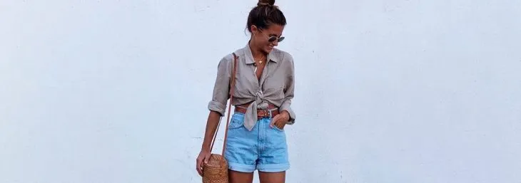 mulher de costas para parede branca usando shorts azul com camisa cinza e óculos de sol levando uma bolsa marrom no ombro